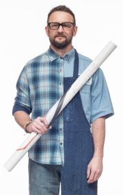 Man with glasses wearing a plaid shirt and denim overalls, holding a rolled blueprint.