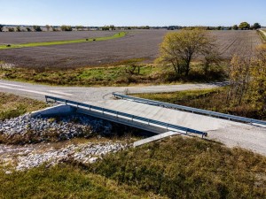 A concrete bridge spanning a gravel road in a rural landscape with fields in the background.
