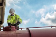 Worker in a safety helmet and high-visibility jacket operating equipment on a large industrial tank.