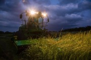 Agricultural equipment cutting grass in a field during twilight hours with headlights on.