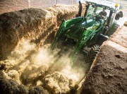 Green tractor operating in a barn, moving hay or straw with a front loader.
