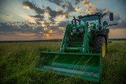 Green tractor with a front loader attachment in a field during sunset with clouds in the sky.