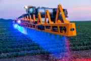 Agricultural sprayer with blue and red LED lights spraying crops in a field at dusk.