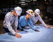 Three workers in hard hats examining a large piece of leather on a table in an industrial setting.