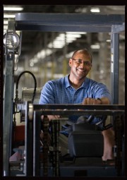 A man sitting in a forklift inside an industrial facility, smiling at the camera.