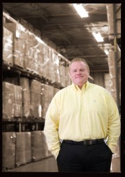 Man wearing a yellow shirt and black pants standing in a warehouse with shelves of boxes in the background.