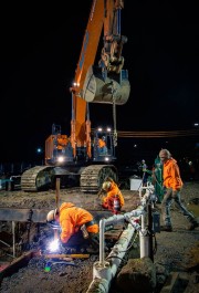 Construction workers in orange jackets operating machinery and welding at a construction site during nighttime.