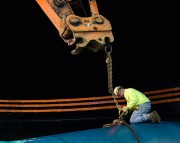 Construction worker in a yellow jacket securing a large pipe with a chain and machinery at night.