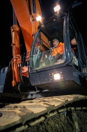 Operator sitting inside an excavator cabin at night with illuminated controls and machinery.