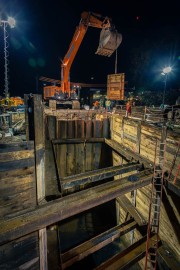 Excavator lifting a large bucket at a construction site during nighttime with bright lights.
