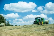 Green tractor working in a field with round hay bales under a blue sky with clouds.