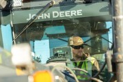 Operator inside a John Deere tractor cab, wearing sunglasses and a cap, with equipment visible in the foreground.