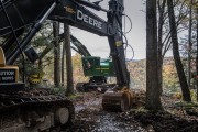 John Deere excavator operating in a forested area with trees and foliage in the background.