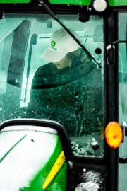 Operator wearing a John Deere cap inside a tractor cabin during snowy weather.