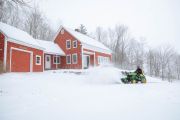 A person using a green tractor to remove snow in front of a red house during a snowstorm.