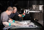 Two workers using a leather press machine in a manufacturing setting, wearing gloves and focused on their task.