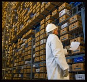 Warehouse worker in a white coat and hard hat checking inventory against a clipboard in a storage facility.