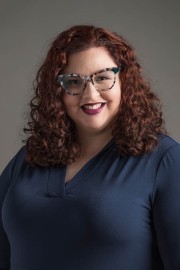 Woman with curly red hair wearing glasses and a blue top, smiling at the camera.