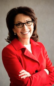 Woman wearing a red blazer and glasses, smiling with arms crossed in a neutral setting.
