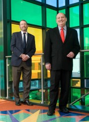 Two men in business suits standing indoors with colorful glass panels in the background.