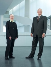 Two individuals standing side by side in formal attire, one wearing a black coat and the other in a dark suit.