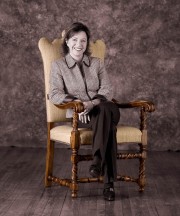A woman in a patterned blazer sitting on a vintage wooden chair with a neutral background.