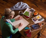 A woman sketching on a blank page at a table surrounded by design materials and a cup of coffee.