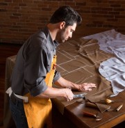 A craftsman using a knife to cut leather material on a table in a workshop setting.
