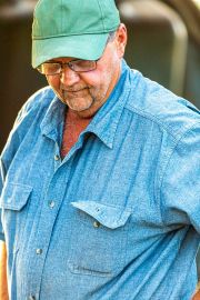 Farmer wearing a blue button-up shirt and a green cap, looking down with glasses on.