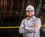 Man wearing a hard hat and safety glasses, standing with arms crossed at an industrial site.