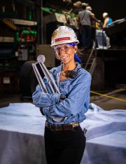 Woman wearing a hard hat and safety glasses, holding measuring tools in a manufacturing environment.
