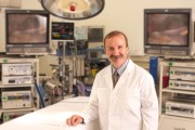 A medical professional wearing a white coat stands in a surgical room with medical equipment in the background.