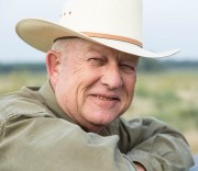 Older Rancher with a cowboy hat smiling while leaning on a wooden fence.