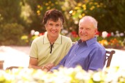 An elderly couple smiling while sitting together on a bench in a garden setting.