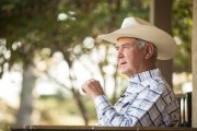 Older Rancher wearing a white cowboy hat and plaid shirt sitting outdoors, engaged in conversation.