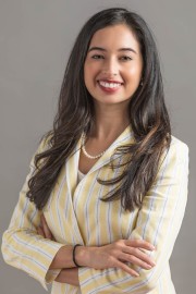 A woman with long dark hair wearing a yellow and white striped blazer and pearl necklace, smiling with arms crossed.
