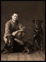 A man kneeling beside two black dogs, one lying down and one sitting, in a studio setting.