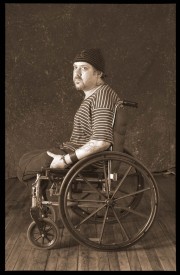 A man sitting in a wheelchair, wearing a striped shirt and a beanie, looking to the side.