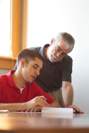A young man in a red shirt writing in a book while an older man observes closely.