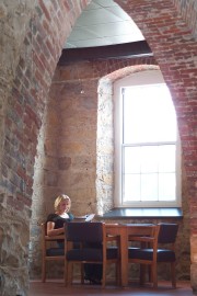 A woman sitting at a wooden table reading a document in a stone-walled room with large windows.