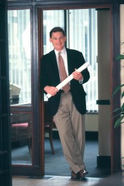 A businessman in a suit holding a rolled blueprint while standing in an office doorway.