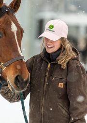 Woman wearing a pink cap and brown jacket interacting with a horse in snowy conditions.