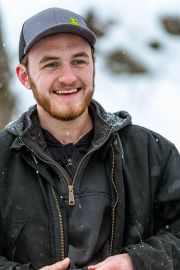 A young man with a beard wearing a black jacket and a gray cap, smiling in a snowy environment.