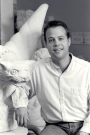 A man smiling while sitting next to a stone sculpture of a creature in a workshop.
