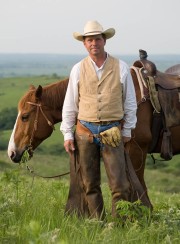 Man wearing cowboy attire stands next to a horse in a rural setting with green grass.