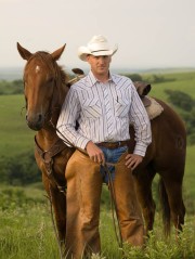 A cowboy wearing a white hat and striped shirt stands next to a brown horse in a grassy field.