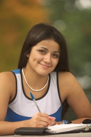 Young woman with long dark hair writing in a notebook while smiling at the camera.