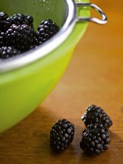 A green colander filled with fresh blackberries, with a few berries placed on a wooden surface.